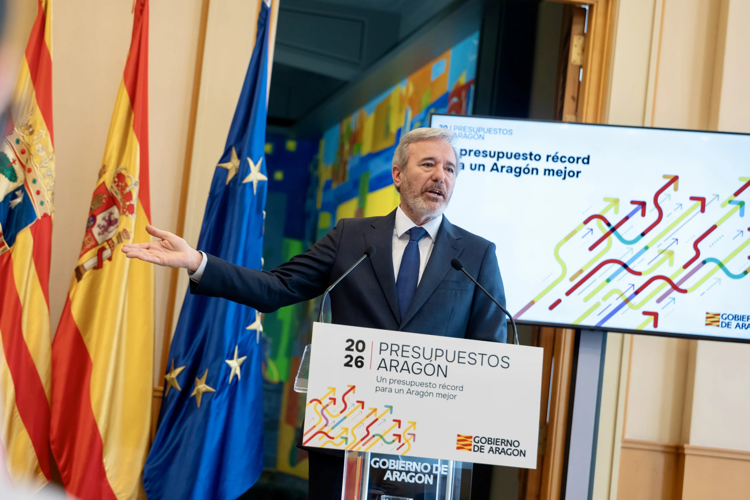 6932d92cbefc6 Jorge Azcçon, presidente del Gobierno de Aragón, presentando el presupuesto 2026 en un atril, con banderas de España, Aragón y la Unión Europea al fondo.